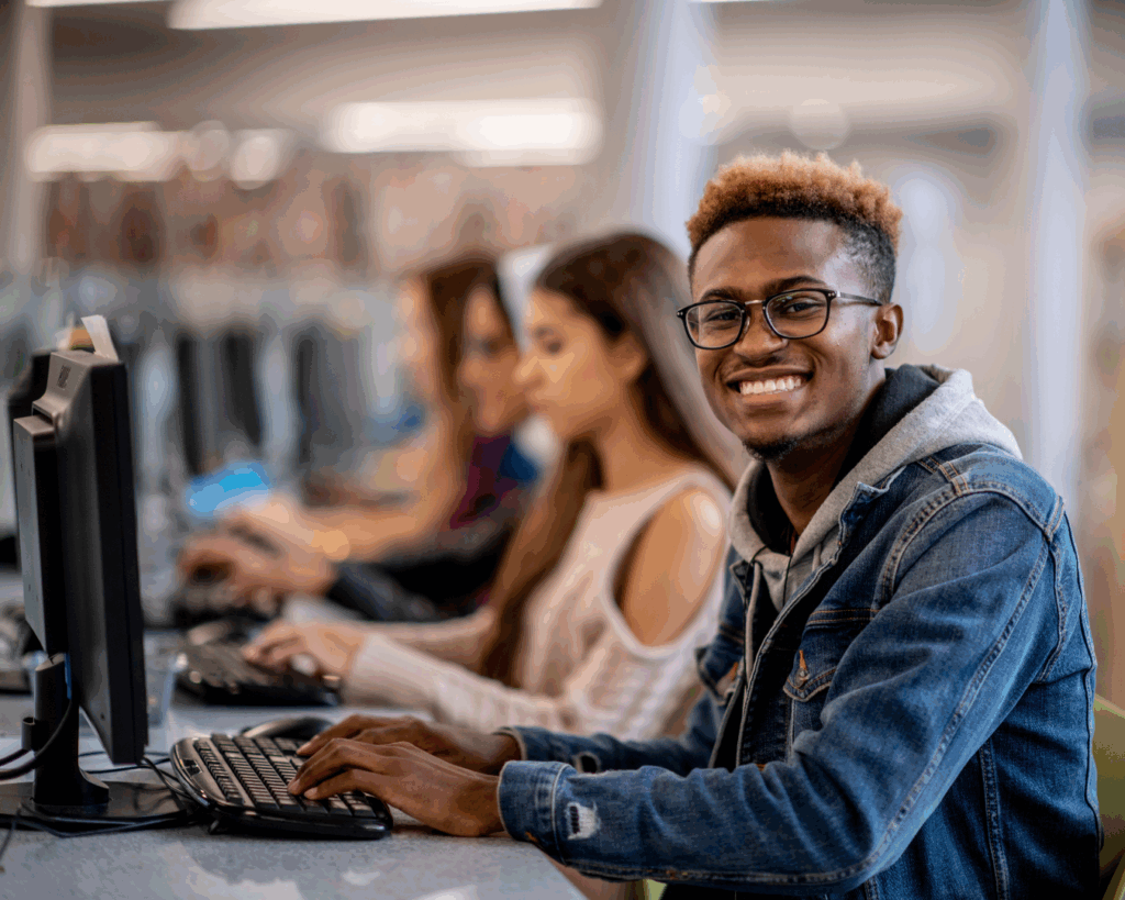 A young man is using a computer in a library or computer classroom. He smiles for a photo.