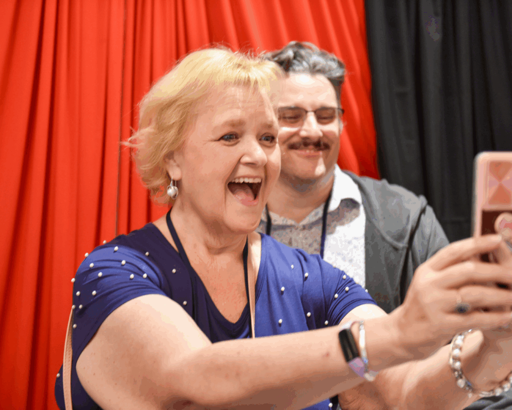 A woman at OLC Accelerate 2024 smiles widely and takes a selfie with another attendee.
